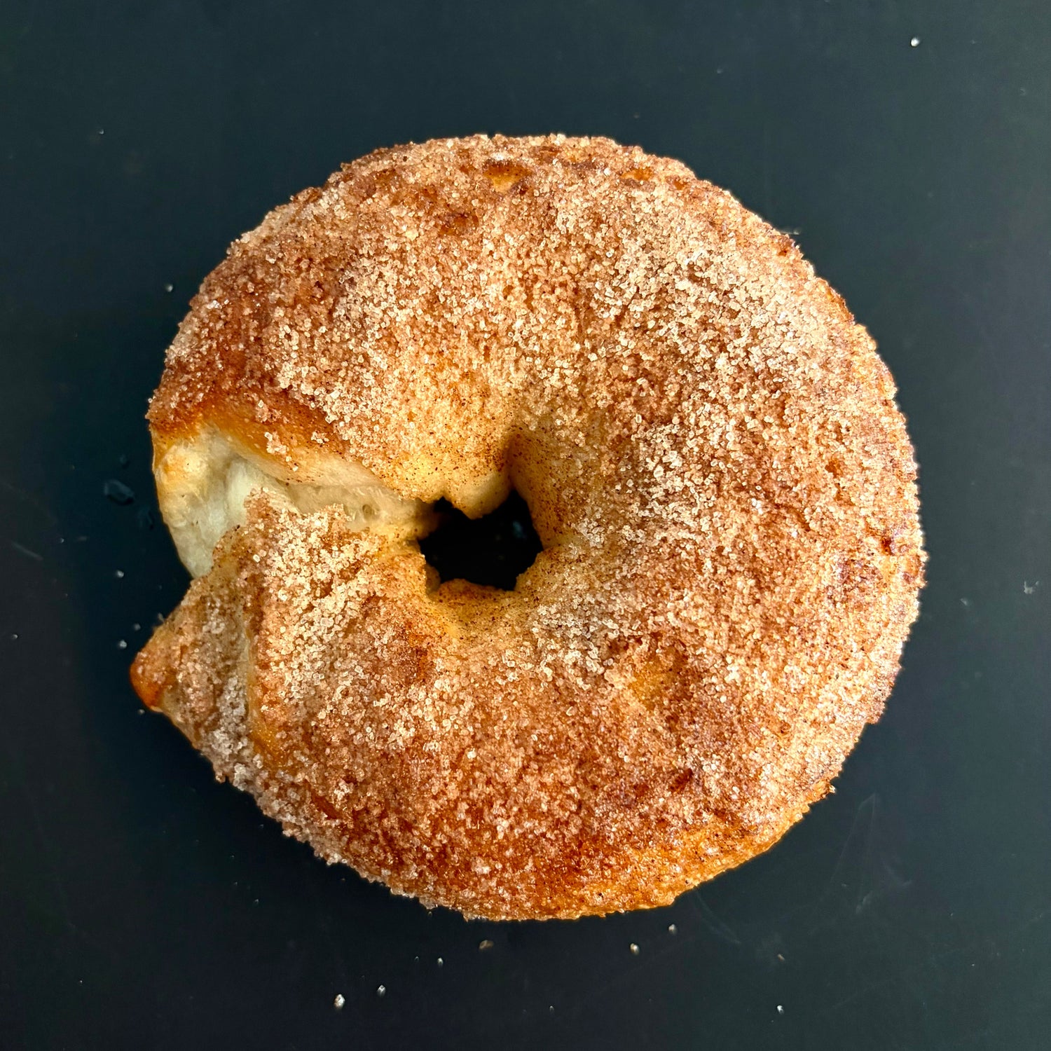 A single cinnamon sugar donut with a bite taken out, resting on a dark black background.