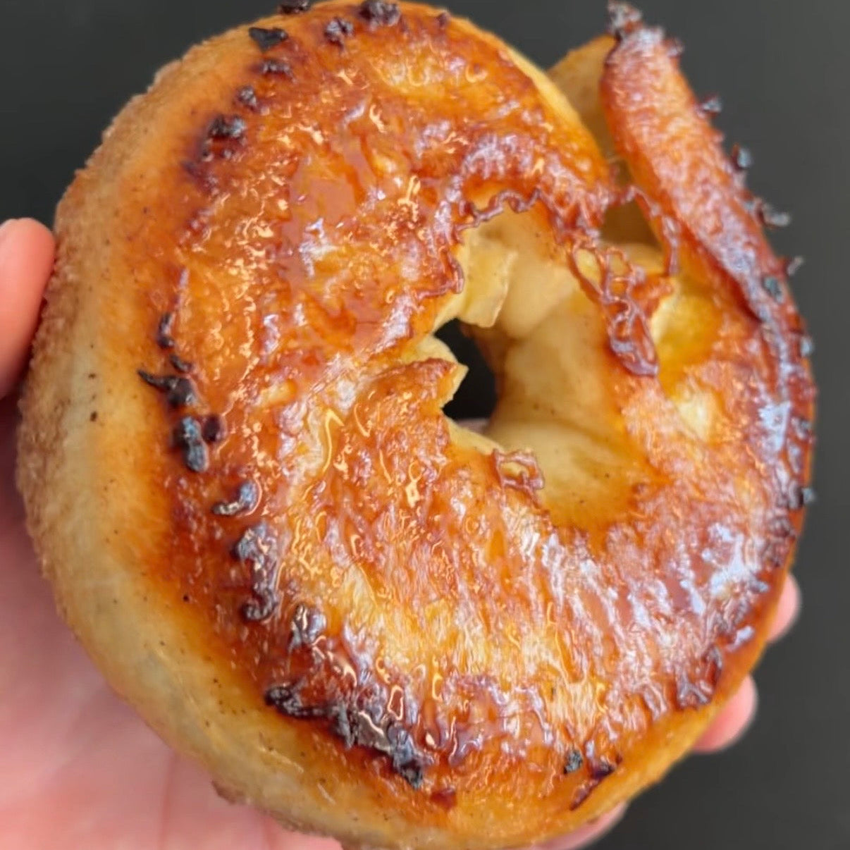 A close-up of a hand holding a glazed bagel with a glossy caramel-like topping against a dark background.